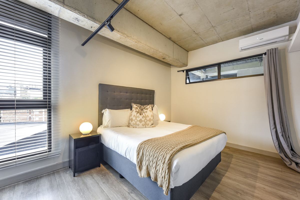 Modern bedroom featuring an exposed concrete ceiling, laminate flooring, split-system air conditioning, and large windows with venetian blinds.