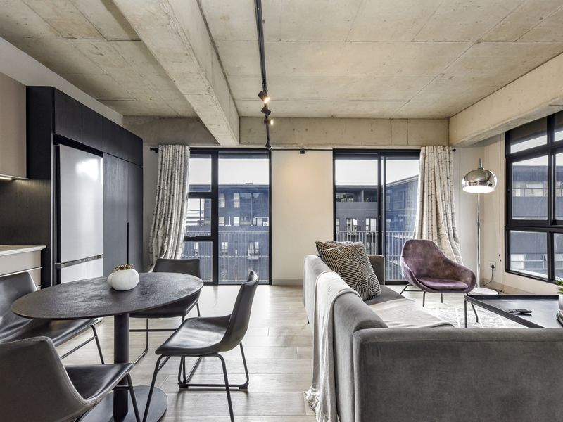Modern open-plan living area featuring an exposed concrete ceiling, track lighting, and sleek built-in cabinetry with large windows providing ample natural light.