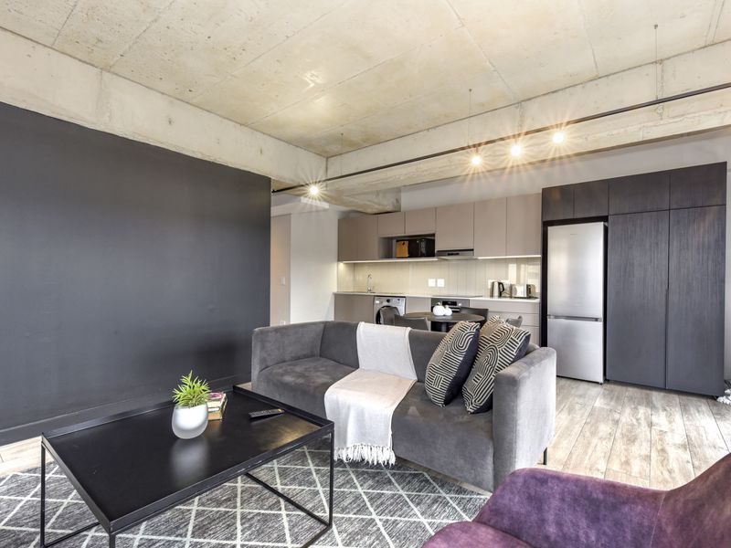 Modern open-plan living area featuring an exposed concrete ceiling, sleek built-in kitchen cabinetry, and high-quality laminate flooring in a contemporary apartment setting.