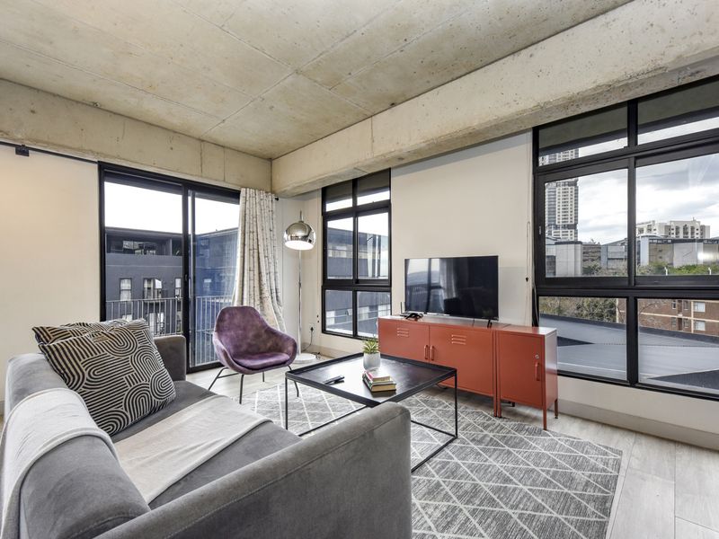 Modern living room featuring an industrial-style exposed concrete ceiling, large black-framed windows, and access to a balcony with urban views.