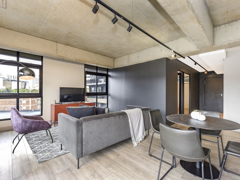 Modern open-plan living and dining area featuring an exposed concrete ceiling, track lighting, and large windows with urban views.