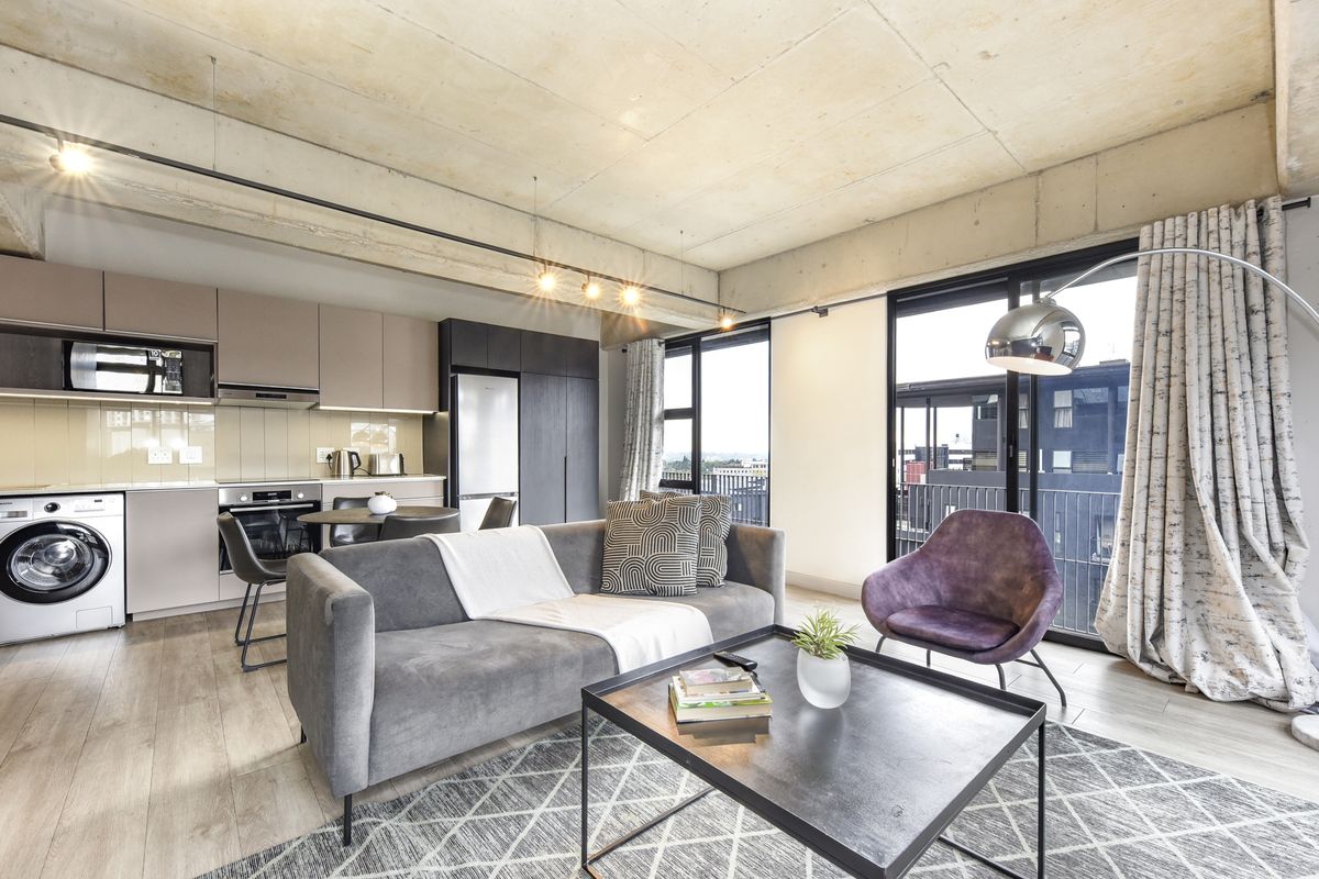 Modern open-plan apartment featuring an exposed concrete ceiling, sleek built-in kitchen cabinetry, integrated appliances, and large sliding doors leading to a balcony.