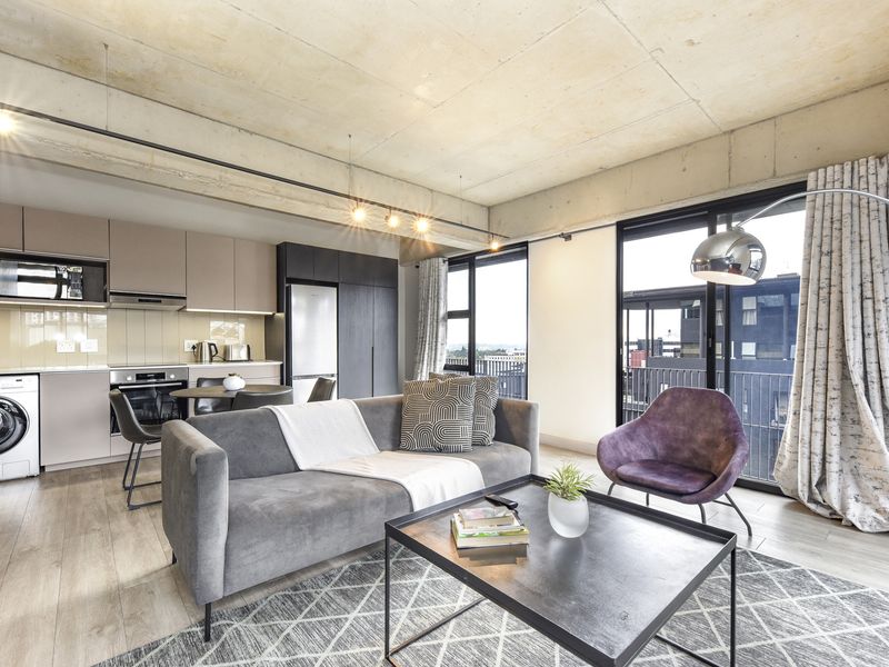 Modern open-plan apartment featuring an exposed concrete ceiling, sleek built-in kitchen cabinetry, integrated appliances, and large sliding doors leading to a balcony.