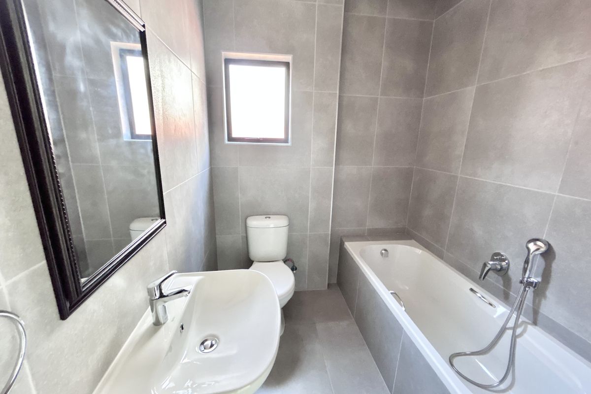A modern, neutral-toned bathroom featuring full-height wall tiling, a built-in bathtub with handheld shower attachment, a pedestal basin, and a dual-flush toilet.