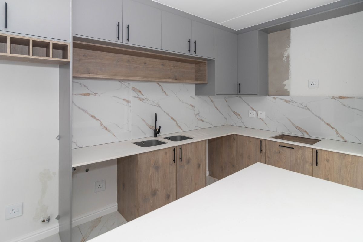 Modern kitchen featuring grey and wood-tone cabinetry, marble-look splashback, stone countertops, and a central island, currently awaiting final wall finishing.