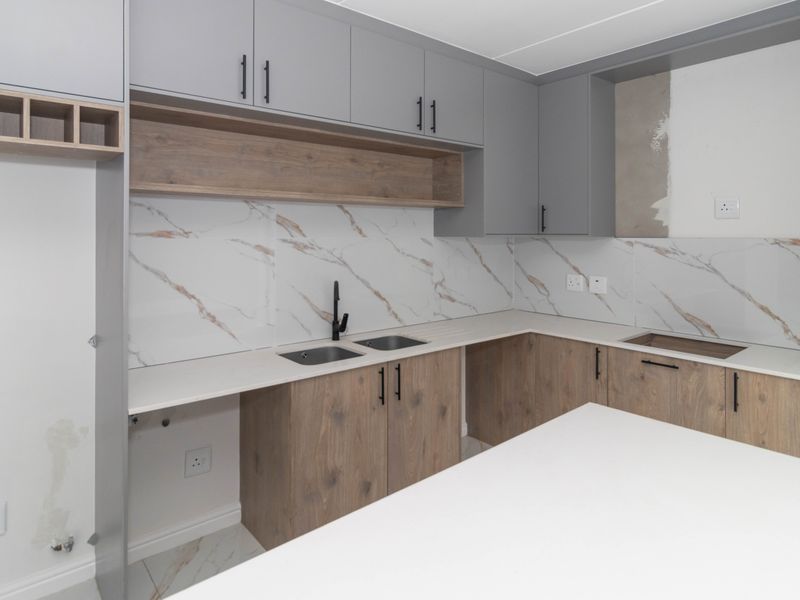 Modern kitchen featuring grey and wood-tone cabinetry, marble-look splashback, stone countertops, and a central island, currently awaiting final wall finishing.