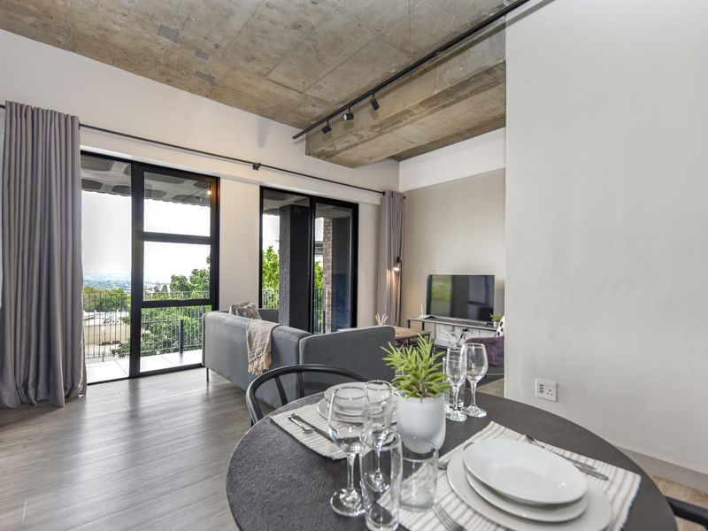 Modern open-plan living area featuring an exposed concrete ceiling, track lighting, and large sliding doors leading to a balcony with scenic views.
