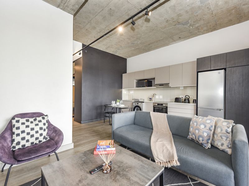 Modern open-plan living area featuring an exposed concrete ceiling, track lighting, and a sleek, well-equipped kitchen with integrated appliances.