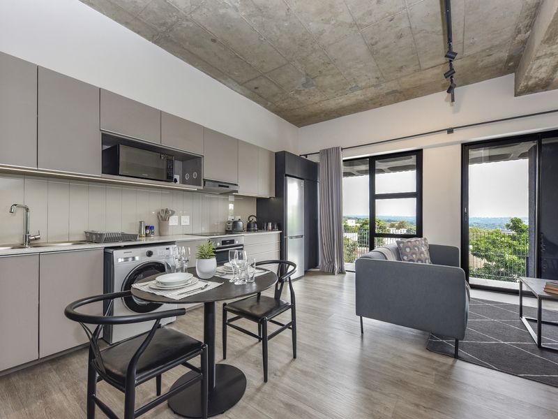 Modern open-plan apartment featuring an exposed concrete ceiling, sleek cabinetry, integrated kitchen appliances, and large sliding doors leading to a balcony with scenic views.