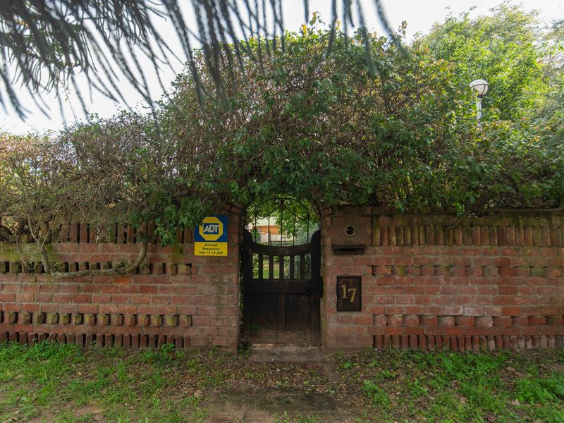 A rustic brick boundary wall featuring a wrought iron pedestrian gate, house number, and security signage, framed by mature overhanging greenery.