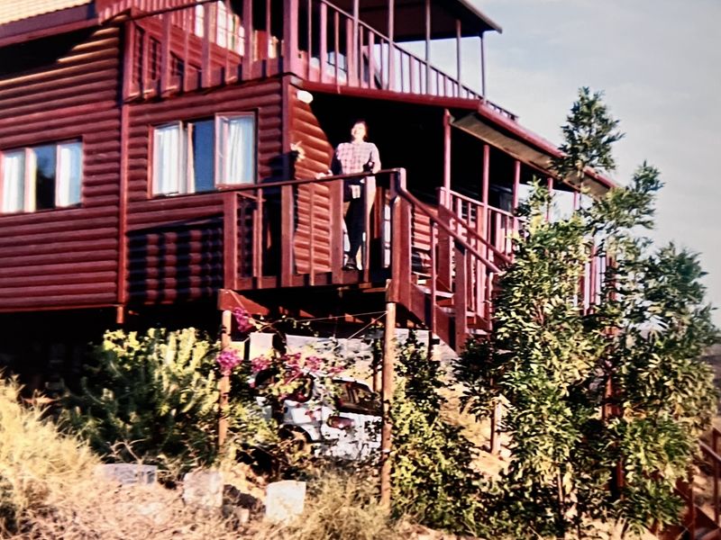 A multi-level timber log cabin featuring a raised deck and balcony, set on a sloped terrain with natural landscaping.