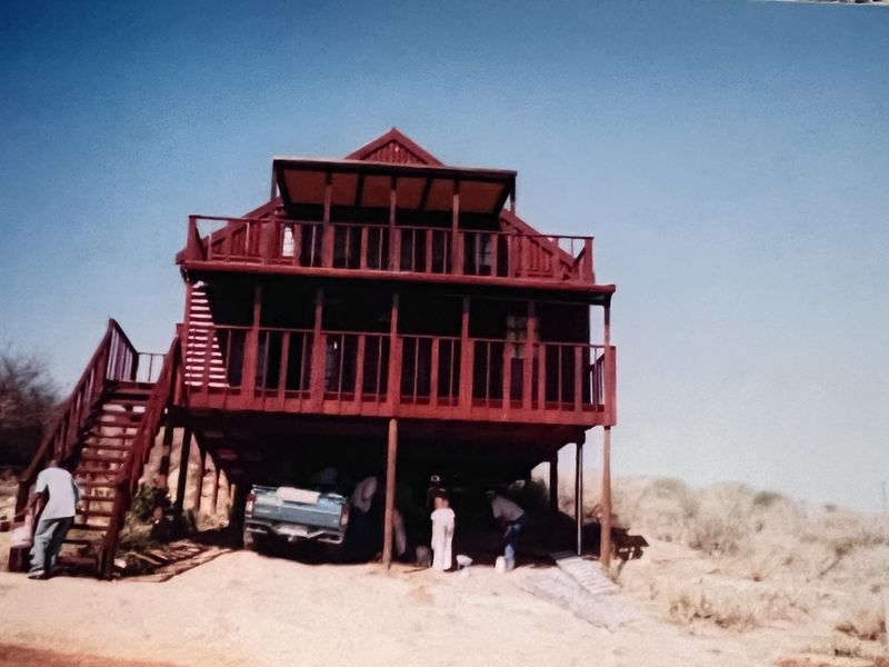 A two-story timber house elevated on stilts, featuring wrap-around balconies and an external staircase, situated in a natural, sandy landscape.