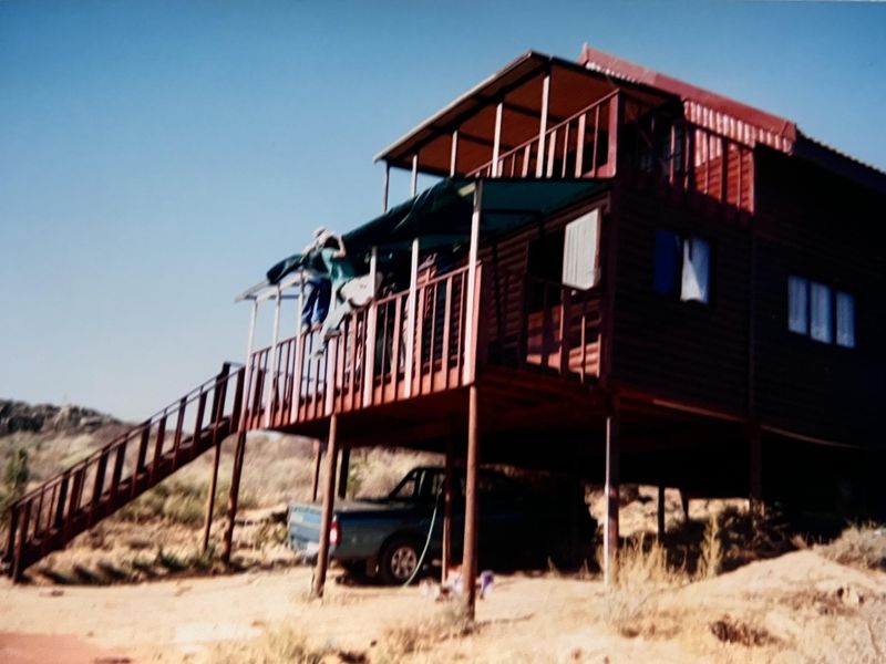 A rustic elevated timber house featuring a wrap-around deck, external staircase, and stilt foundation, set in a natural landscape.