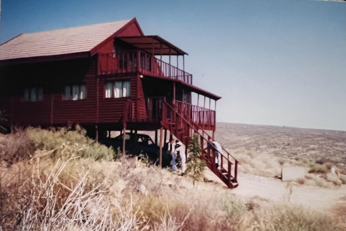 A raised timber-clad house featuring a balcony and external staircase, situated on a sloped landscape with a rural outlook.