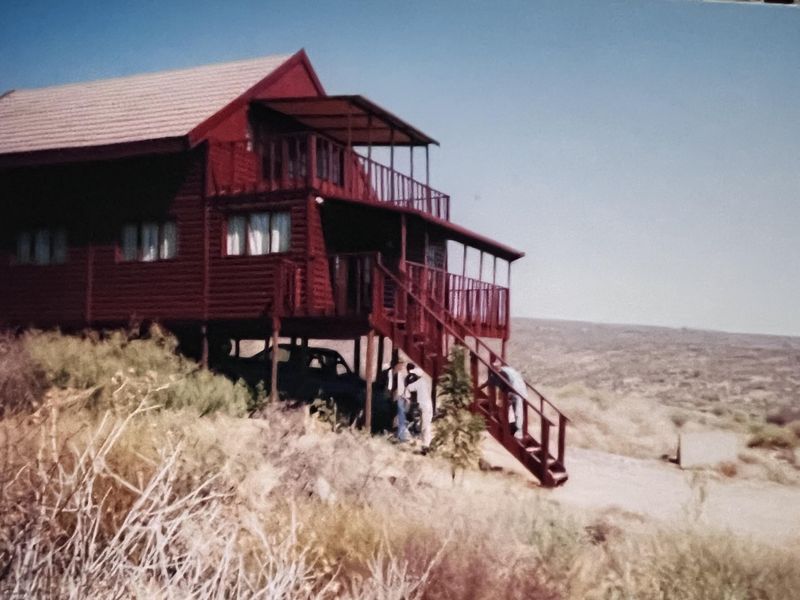 A raised timber-clad house featuring a balcony and external staircase, situated on a sloped landscape with a rural outlook.