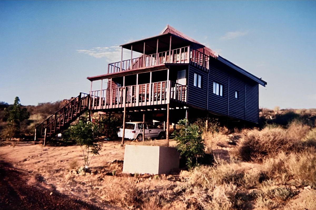 Elevated timber-clad house featuring a wrap-around deck, external staircase, and corrugated metal roofing, situated on a natural, rugged landscape.