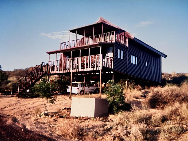 Elevated timber-clad house featuring a wrap-around deck, external staircase, and corrugated metal roofing, situated on a natural, rugged landscape.