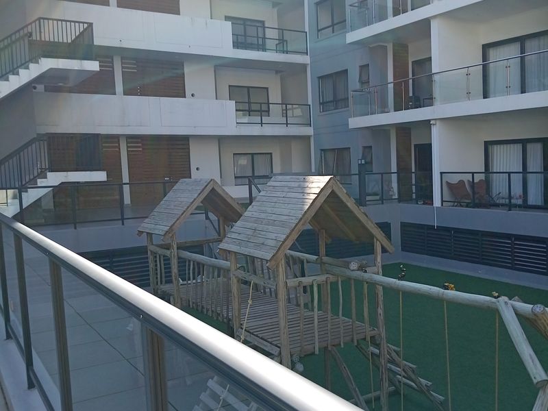 View of an apartment complex courtyard featuring a glass-railed balcony, tiled patio area, and a communal artificial lawn with a wooden play structure.