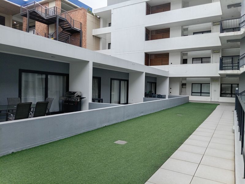 A modern apartment complex exterior featuring a shared artificial turf courtyard, paved walkways, and covered patio areas with sliding door access.