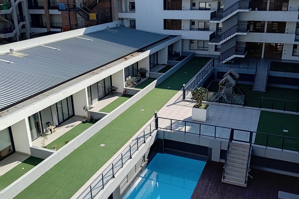 Aerial view of an apartment complex courtyard featuring a swimming pool, timber decking, artificial lawn areas, and modern building architecture.