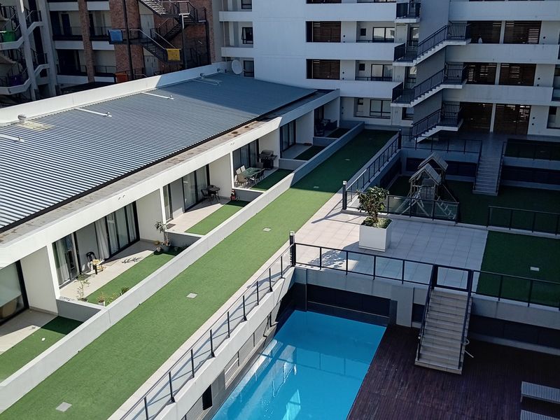 Aerial view of an apartment complex courtyard featuring a swimming pool, timber decking, artificial lawn areas, and modern building architecture.