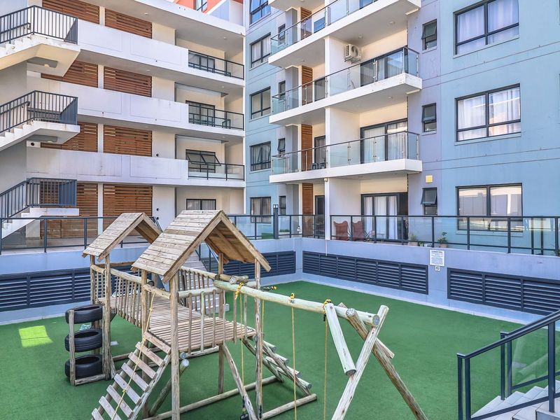 A well-maintained communal courtyard area featuring artificial turf, a wooden playground structure, and modern apartment building facades with glass-fronted balconies.