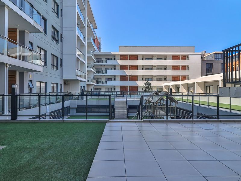 A view of the apartment complex courtyard featuring a tiled patio area, artificial lawn, and modern multi-story building facades with glass balustrades.