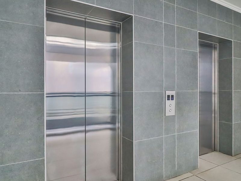 A modern apartment building hallway featuring dual stainless steel elevator doors and contemporary grey tiled wall finishes.