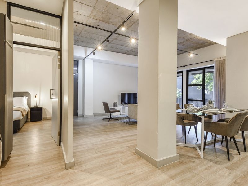 Modern open-plan apartment featuring exposed concrete ceilings, track lighting, and light wood-look flooring, seamlessly connecting the living, dining, and kitchen areas.