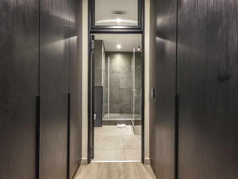 A modern hallway featuring dark built-in cabinetry leading into a contemporary bathroom with tiled finishes and a glass shower enclosure.