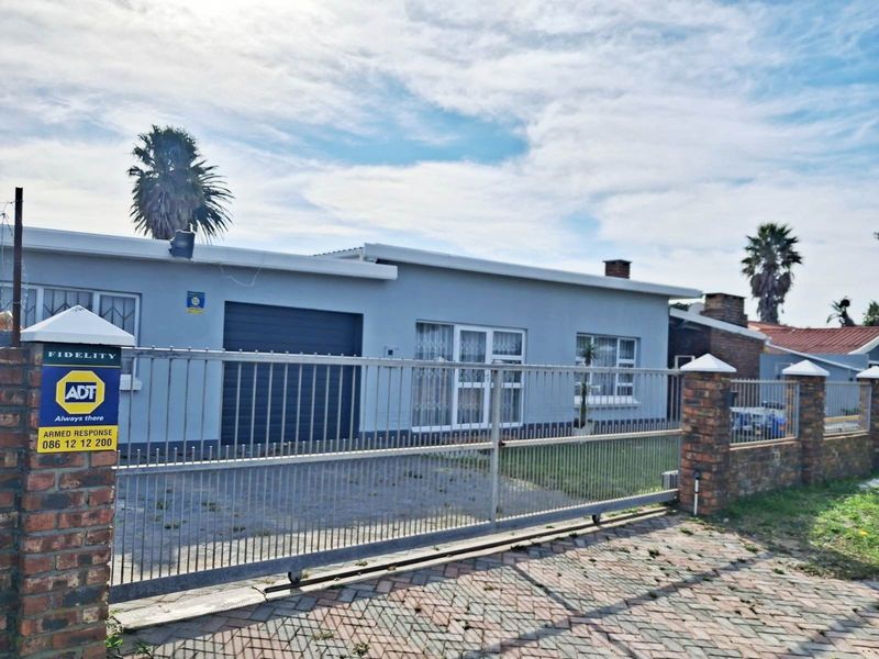 Front exterior view of a single-story house featuring a paved driveway, secure metal gate, brick boundary wall, and a garage.