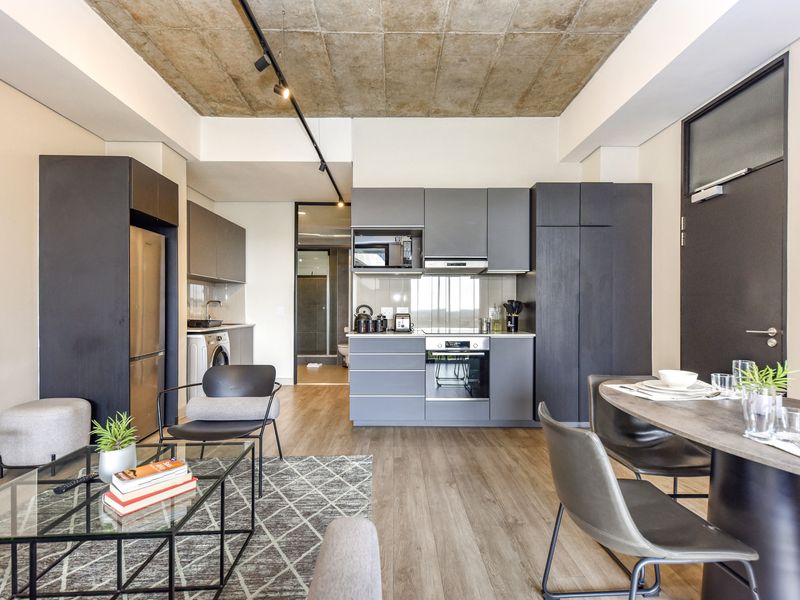 Modern open-plan apartment featuring a sleek kitchen with built-in appliances, exposed concrete ceiling, track lighting, and durable laminate flooring throughout the living space.