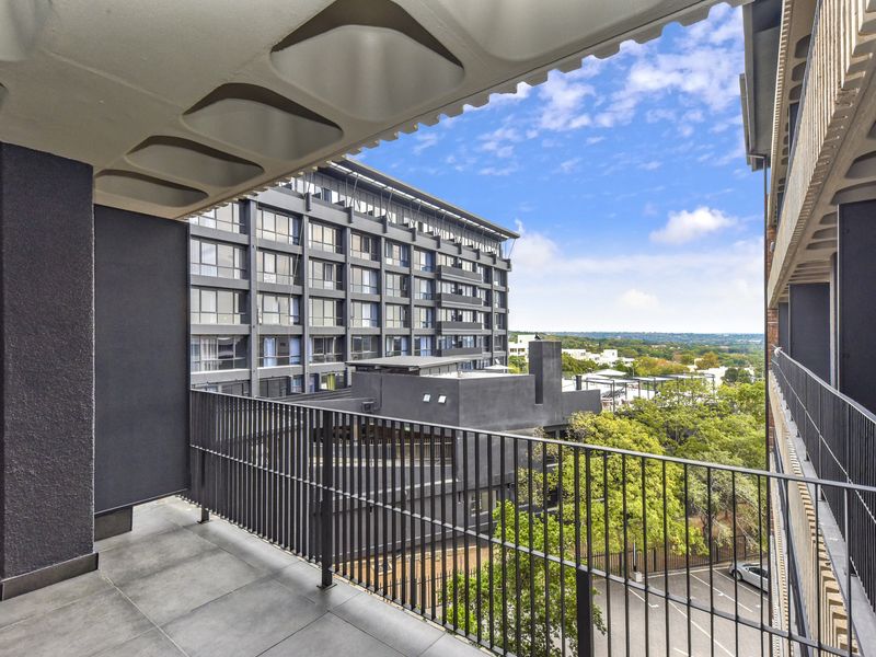 A modern, covered balcony featuring tiled flooring, a sleek metal balustrade, and expansive views of the surrounding urban landscape.