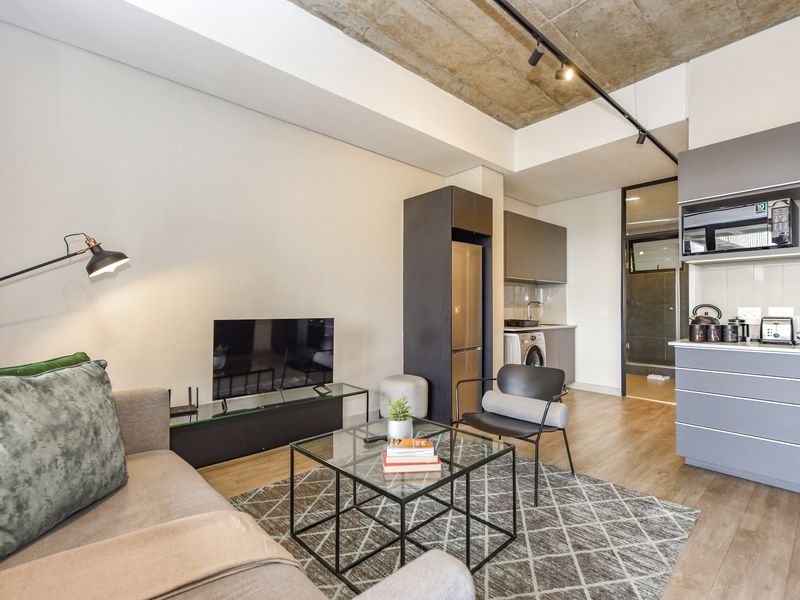 Modern open-plan living area featuring an exposed concrete ceiling, track lighting, and a sleek, contemporary kitchen with integrated cabinetry and high-quality finishes.