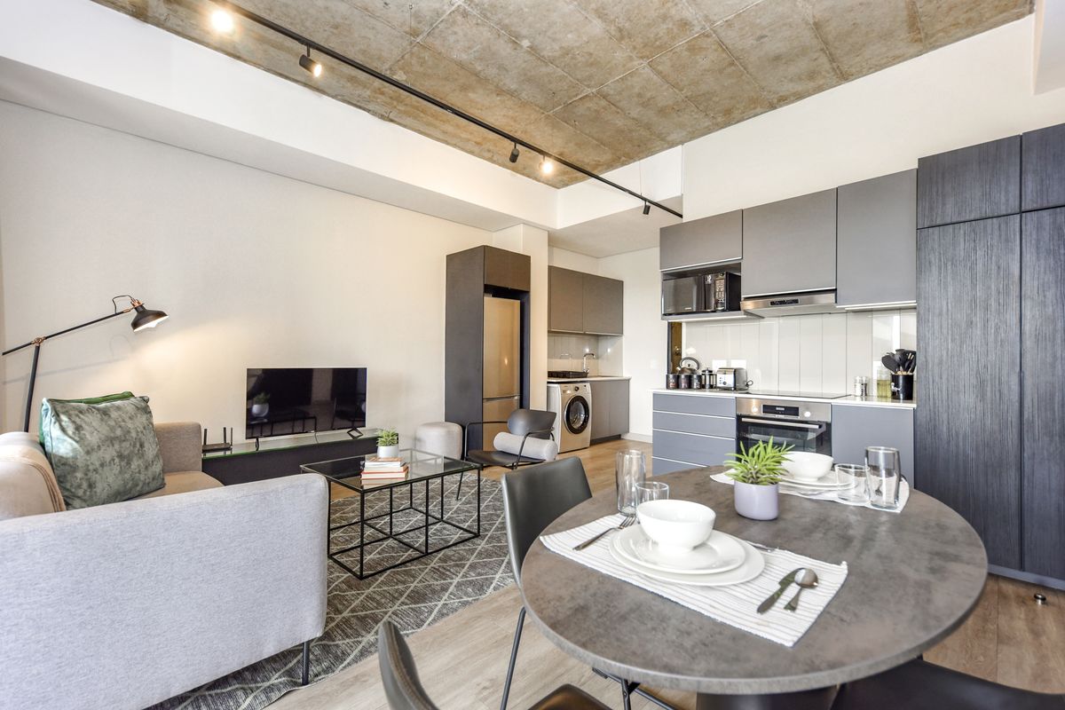 Modern open-plan living area featuring an exposed concrete ceiling, track lighting, and a sleek, fully equipped kitchen with integrated appliances.