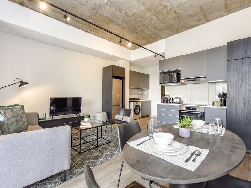 Modern open-plan living area featuring an exposed concrete ceiling, track lighting, and a sleek, fully equipped kitchen with integrated appliances.