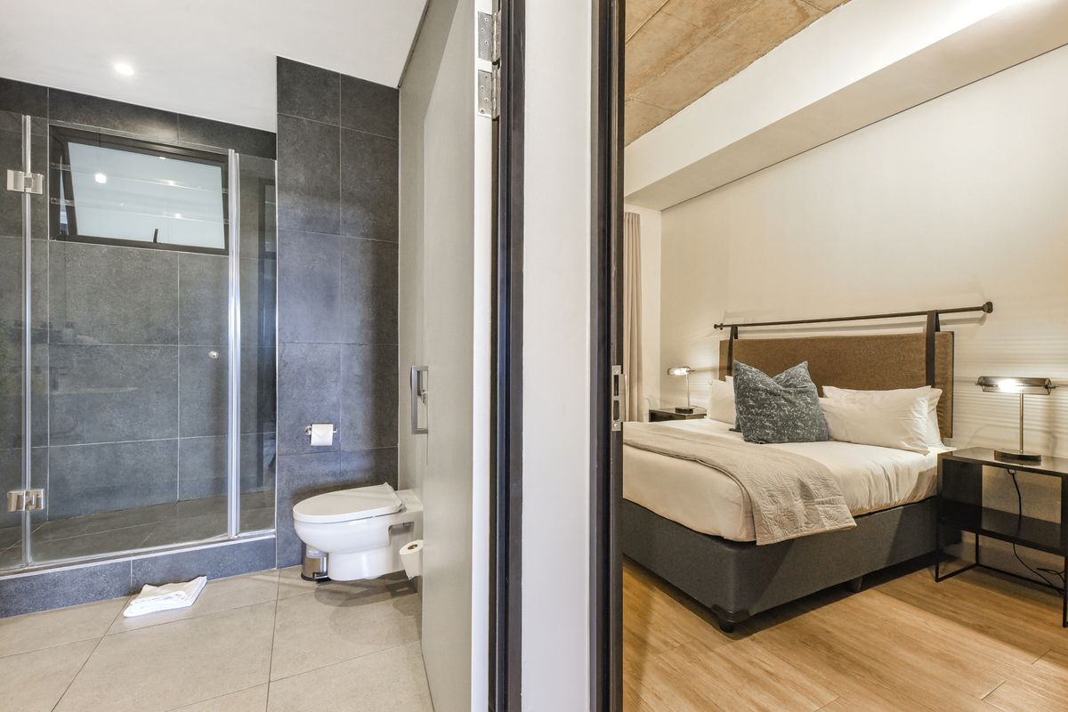 Modern bedroom featuring an en-suite bathroom with a glass shower enclosure, wall-mounted toilet, and contemporary finishes with exposed concrete ceiling details.