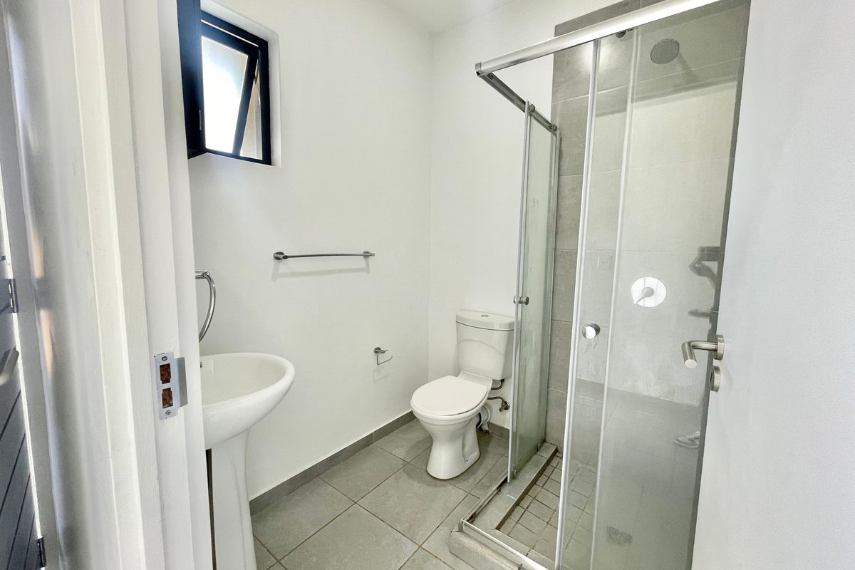 A clean, modern bathroom featuring a pedestal basin, a toilet, and a glass-enclosed shower with neutral floor tiling.