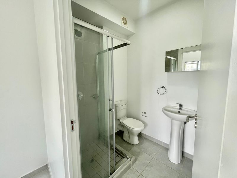 A clean, functional bathroom featuring a pedestal basin, a glass-enclosed shower, and a standard toilet with neutral floor tiling.