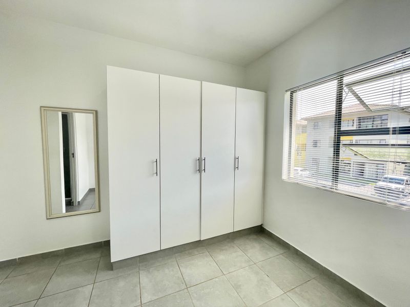 A clean, empty bedroom featuring neutral tiled flooring, built-in white cupboards, and a window with fitted venetian blinds.
