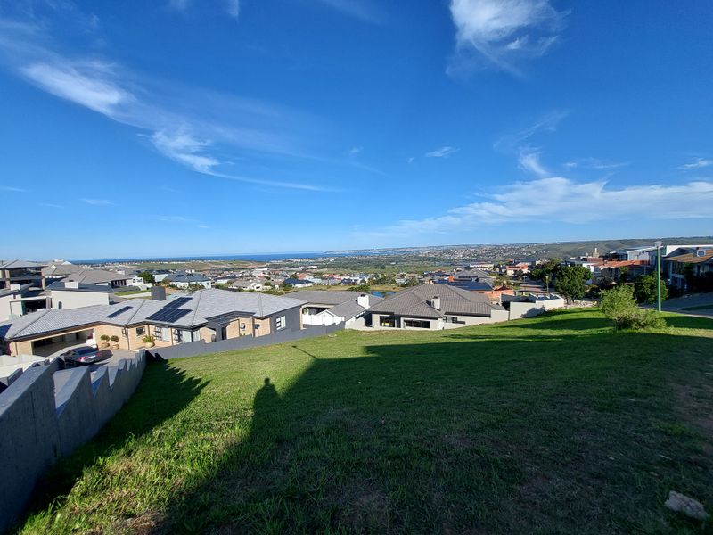 A wide-angle view overlooking a residential neighborhood and distant coastline from a sloped vacant residential plot.