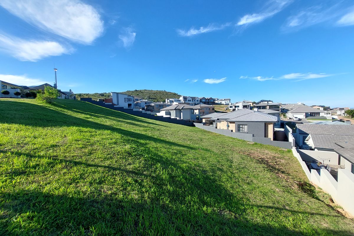 A view of a sloped residential plot overlooking a developed neighborhood with established homes and retaining walls.