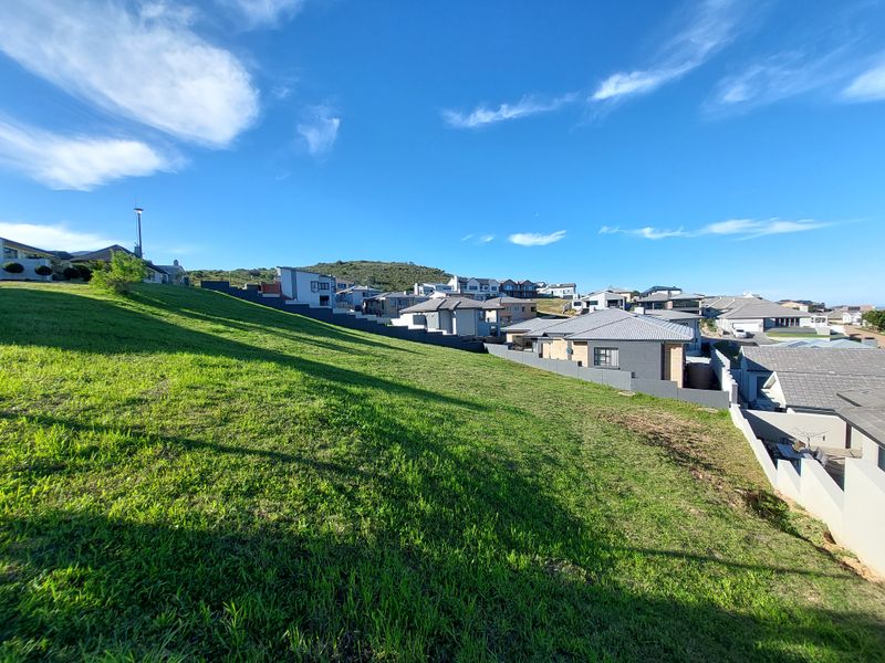 A view of a sloped residential plot overlooking a developed neighborhood with established homes and retaining walls.