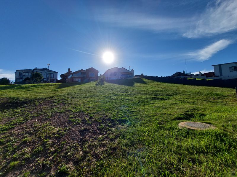 A view of a vacant residential plot with a gentle slope, grass cover, and a visible manhole cover, set against a clear blue sky.