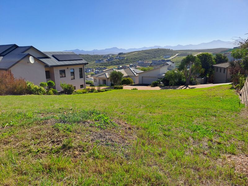 A view of the vacant residential land featuring sloped grass terrain with surrounding neighborhood homes and distant mountain scenery.