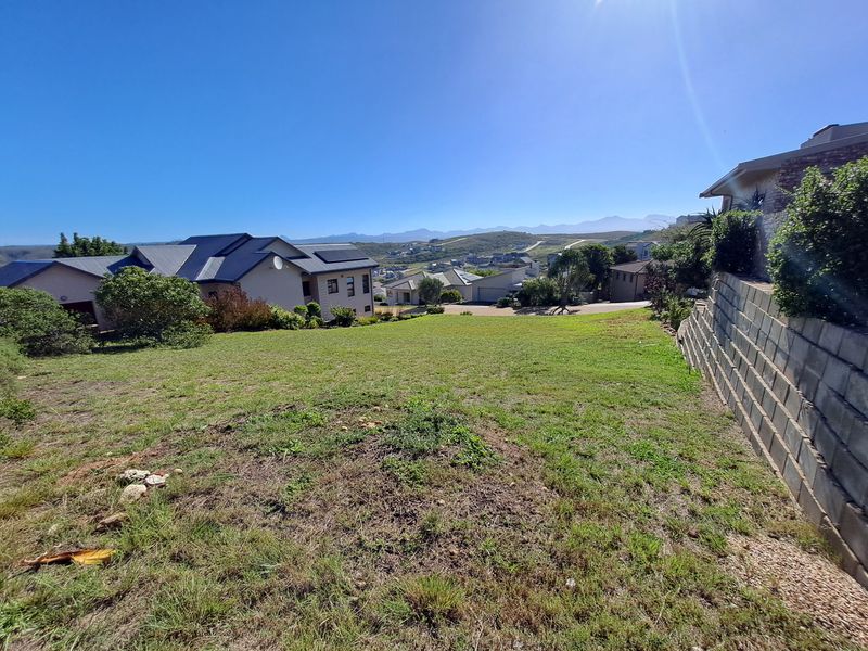 A sloped residential vacant land plot featuring a concrete block retaining wall and views of the surrounding neighborhood and distant mountains.