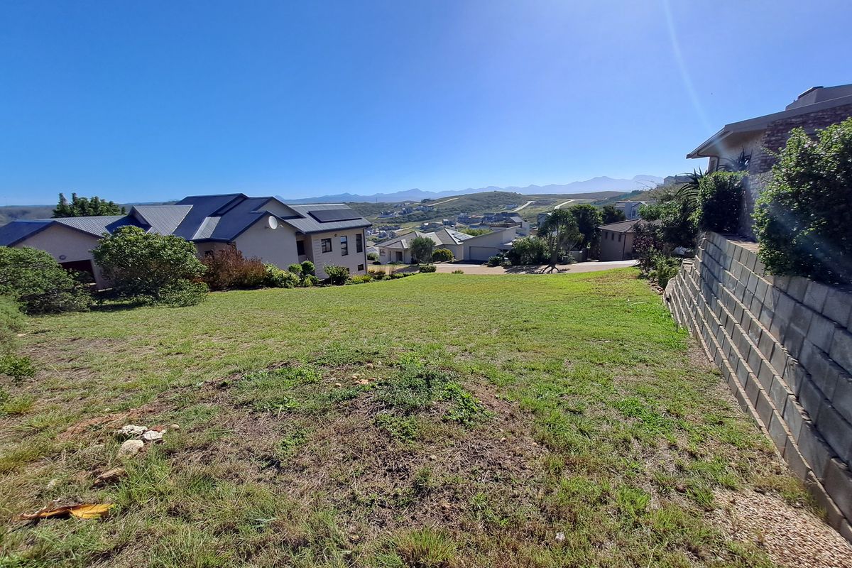 A sloped residential vacant land plot featuring a concrete block retaining wall and views of the surrounding neighborhood and distant mountains.