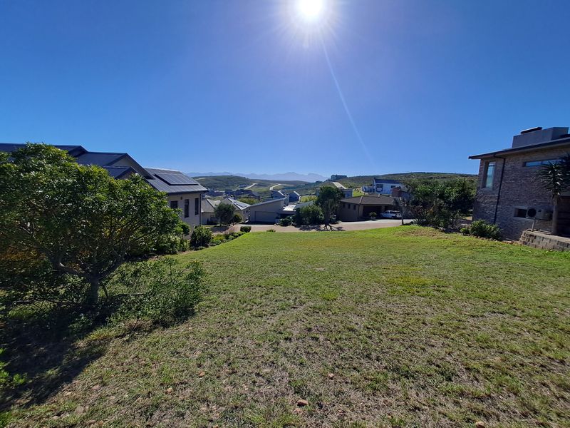 A wide-angle view of a residential vacant land plot featuring a gentle slope and established grass, overlooking the surrounding estate and distant mountain range.