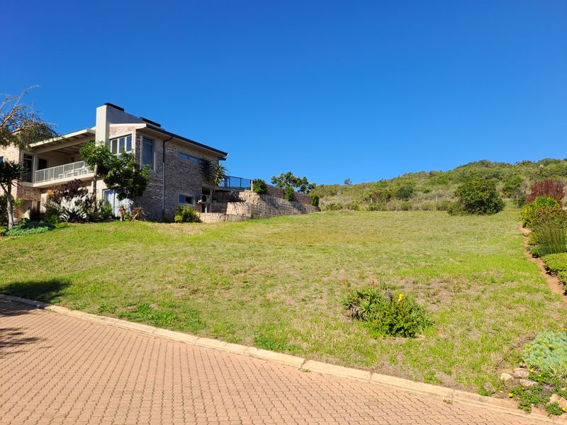 A view of a residential plot featuring a paved driveway, stone retaining walls, and established landscaping adjacent to a multi-story stone-clad home.