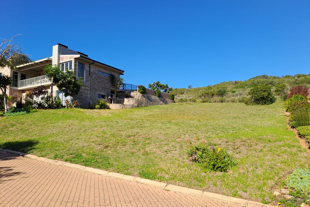 A view of a residential plot featuring a paved driveway, stone retaining walls, and established landscaping adjacent to a multi-story stone-clad home.